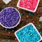 Three bowls: one filled with blue sprinkles, one with pink sprinkles, one with purple sprinkles