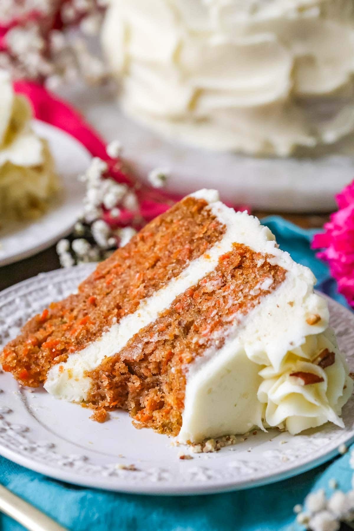 A slice of the best carrot cake recipe on a white plate with white flowers in the background. A bite missing from the slice.