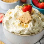 Bowl of cheesecake dip with graham cracker crumbs, a graham cracker, and a strawberry on top.