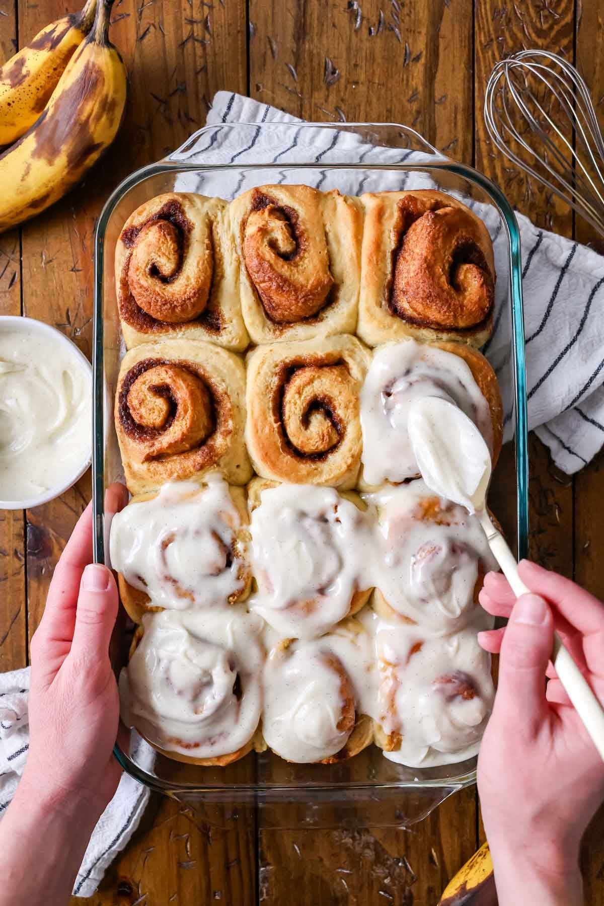 Overhead view of banana bread cinnamon rolls being frosted with cream cheese icing.