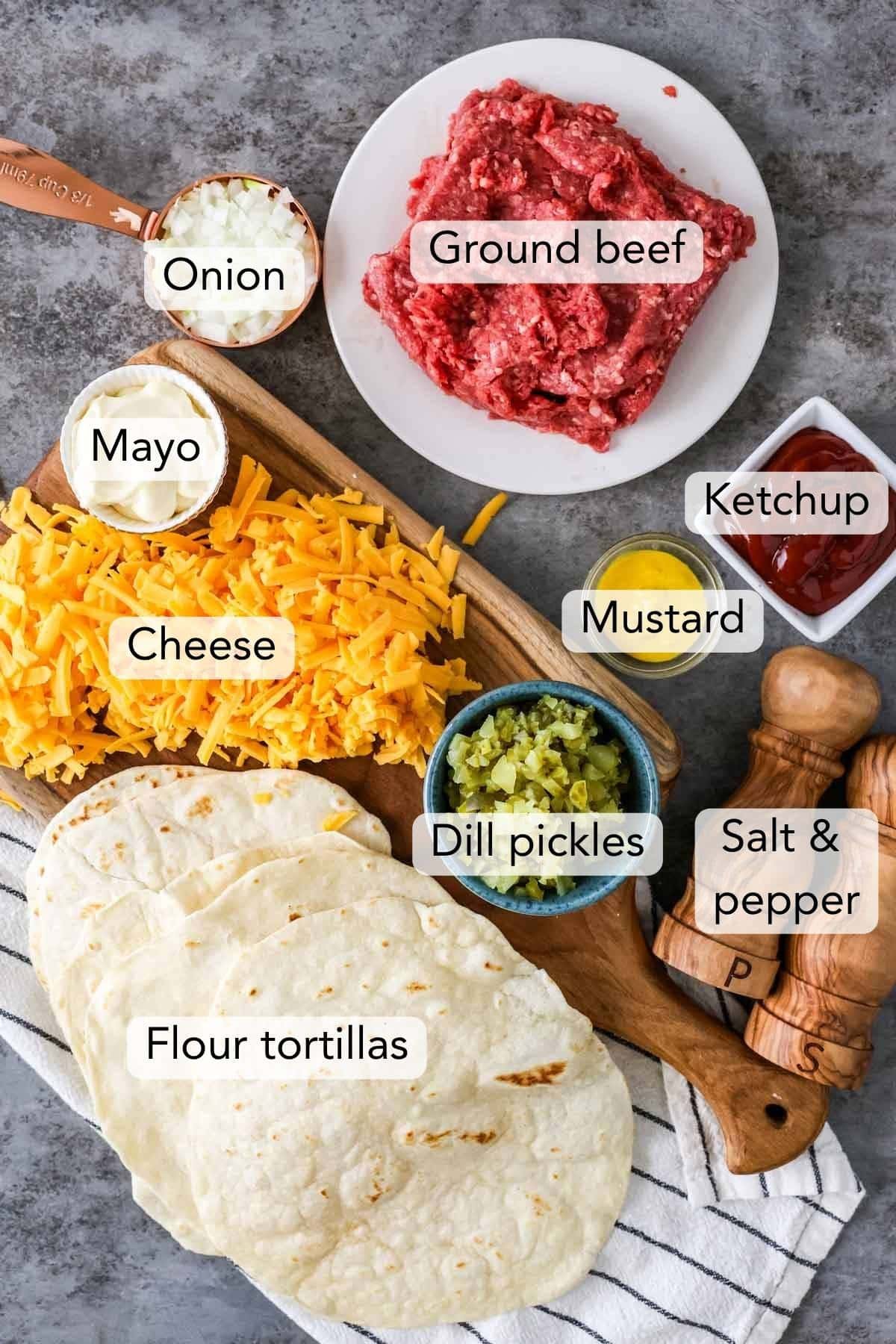 Overhead view of ingredients for cheeseburger tortillas including ketchup, ground beef, pickles, tortillas, and more.